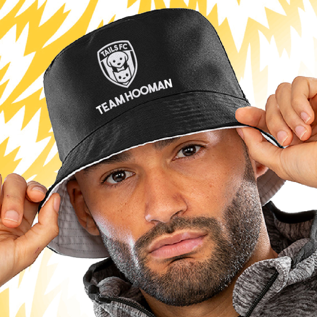 Man wearing a black bucket hat with 'TAILS FC' and 'TEAM HOOMAN' text against a yellow and white background