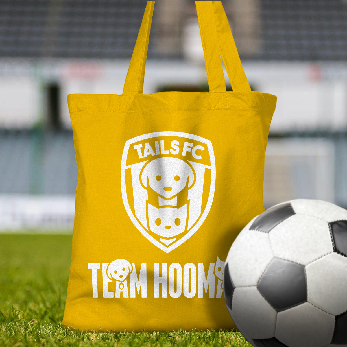 Yellow tote bag with 'TAILS FC' and 'Team Hooman' design on a grassy field with a soccer ball.